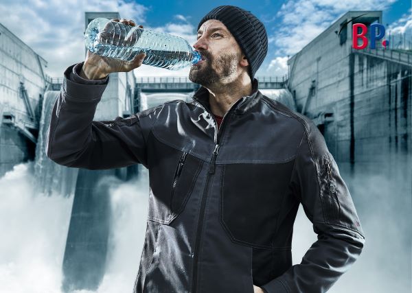 Mann in schwarzer Arbeitskleidung trinkt Wasser vor einem Wasserstaudamm.