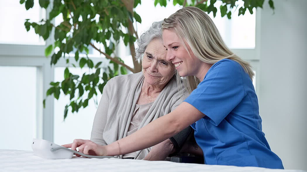 Seniorenpflegerin betreut eine Seniorin und trägt einen Kasack.