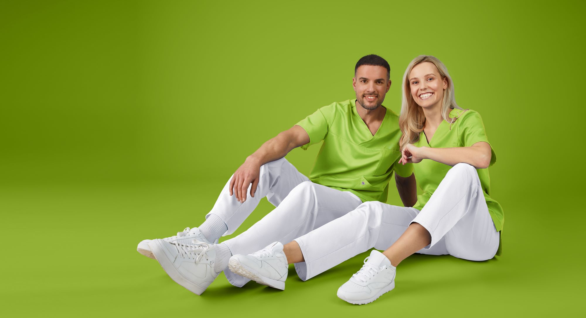 Mann und Frau, beide in grünem Tencel-Kasack und weißer Hose, sitzen auf dem Boden.