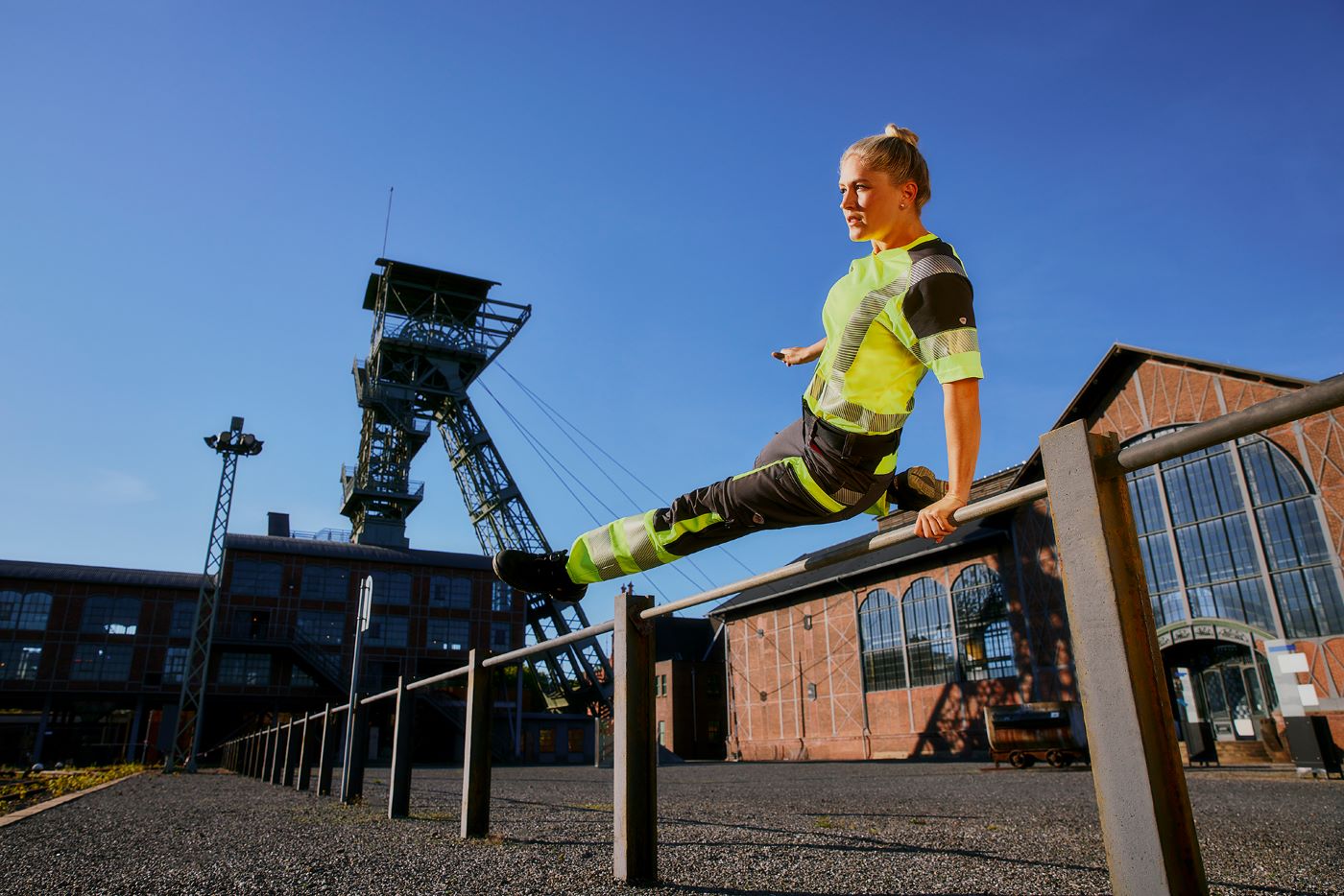 Frau in neongelber und flexibler Arbeitskleidung springt über eine Stange.