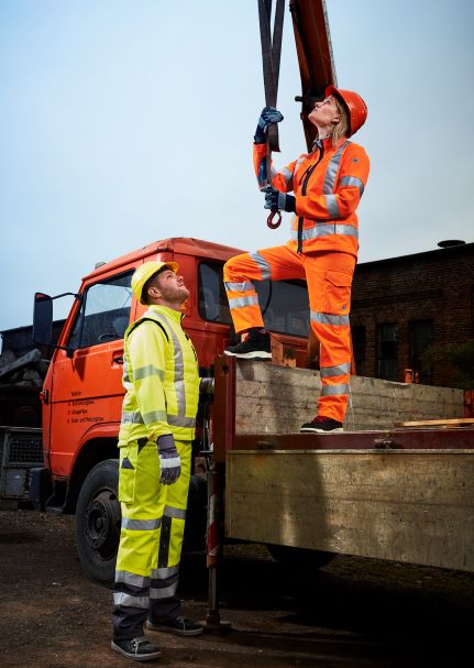 A woman and a man on a construction site are wearing yellow and orange high-visibility clothing.