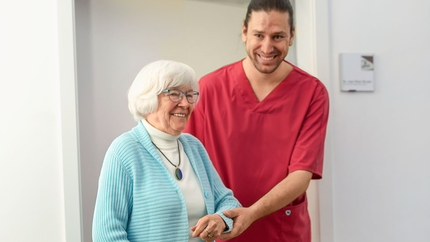 Ouderenverzorger in een BP-jas helpt een oudere te lopen.