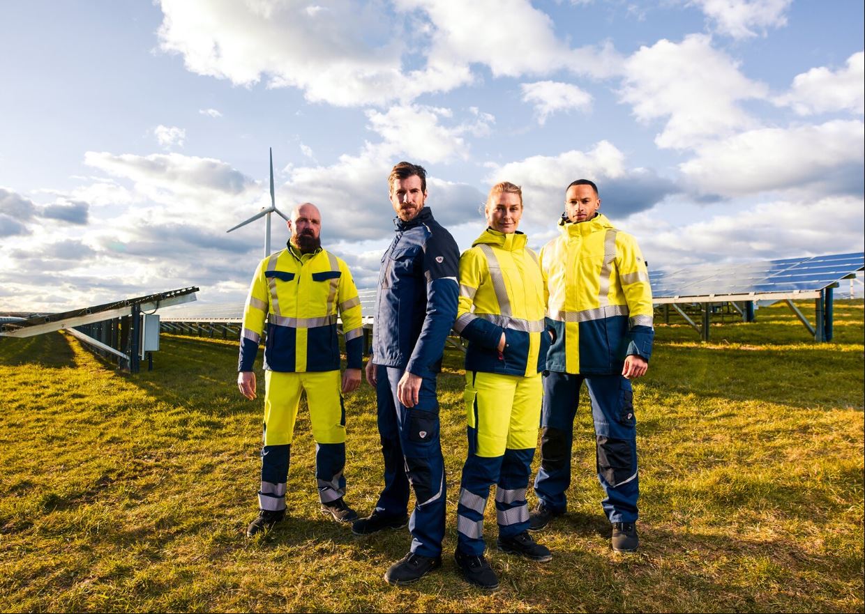 Quatre personnes portant des vêtements de travail modernes à haute visibilité devant des installations d'énergie solaire et éolienne.