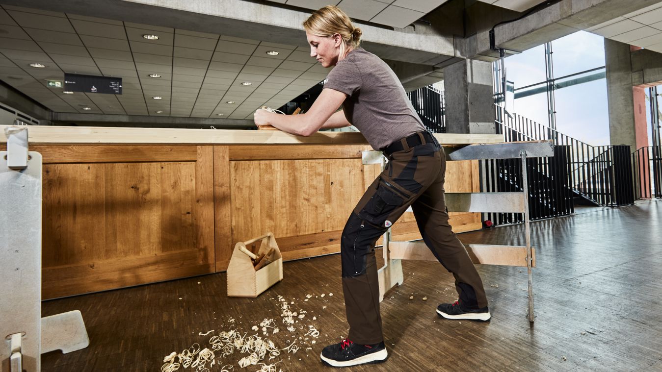 Schreinerin trägt braune Arbeitshose und schleift Holz.