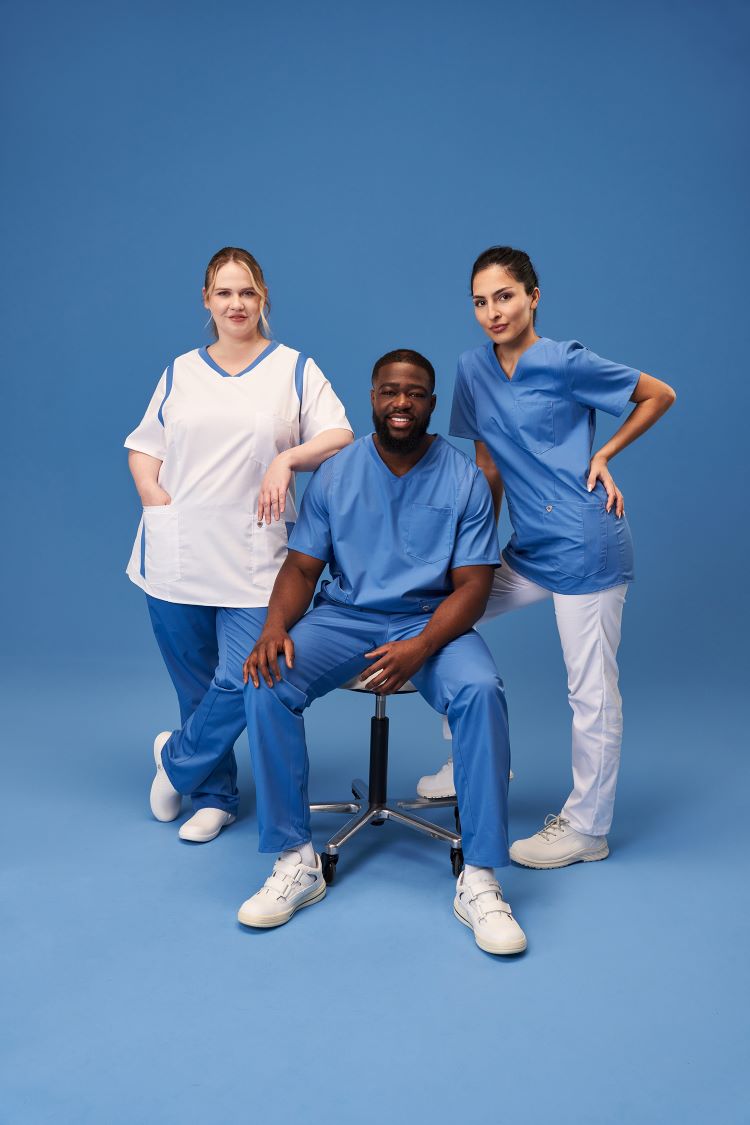 Zwei Frauen und ein Mann tragen medizinische Arbeitskleidung. Der Mann sitzt auf einem Hocker.