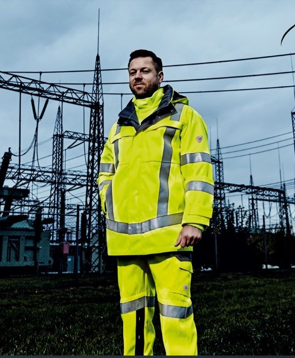 Een energiemanager draagt neongeel werkkleding en staat voor een onderstation.