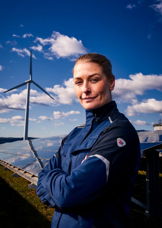 Eine Frau mit eingeschränkten trägt Multinorm Schutzkleidung. Im Hintergrund ist eine Windkraftanlage und Solaranlagen zu sehen.