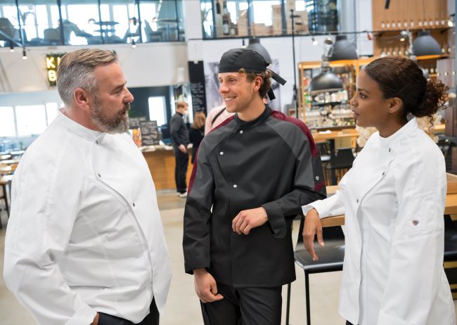 Zwei Männer und eine Frau tragen in einem Restaurantlokal Gastronomiekleidung.