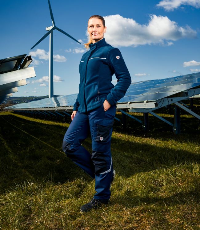 Eine Frau trägt Arbeitskleidung. Hinter Ihr stehen Solaranlagen.