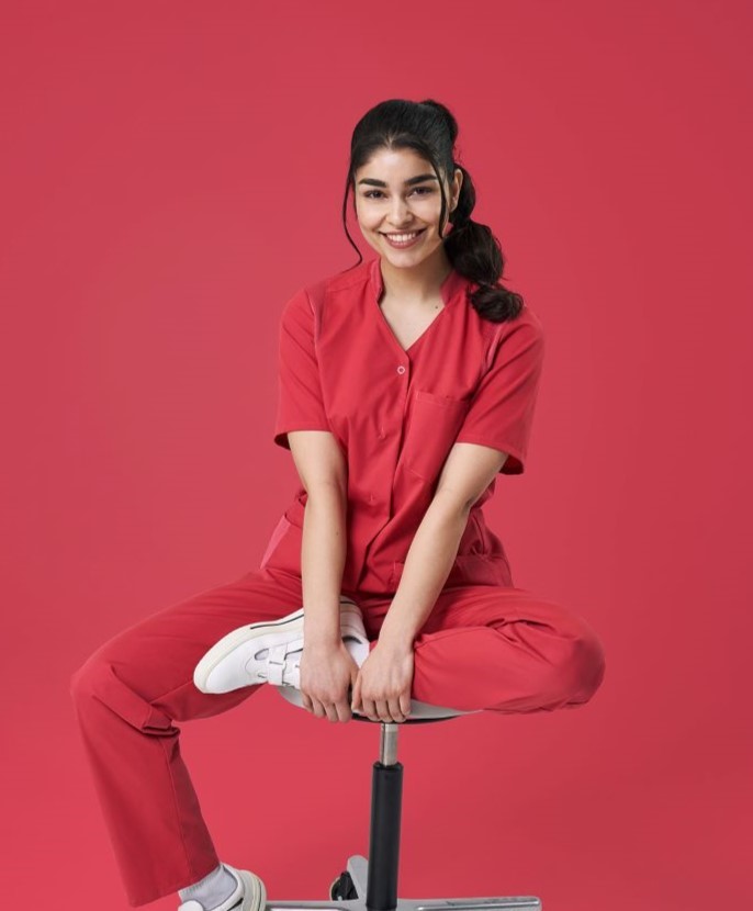 Une aide-soignante porte des vêtements de travail de couleur corail et est assise sur un tabouret.