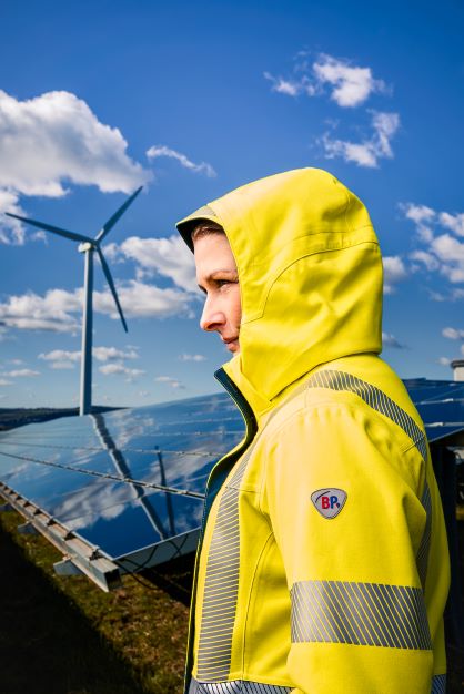 Eine Frau trägt eine Multinormschutzkleidung mit aufgezogener Kapuze in neongelb. Im Hintergrund ist eine Solaranlage.