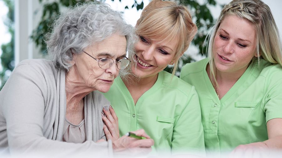 Pflegerinnen in grünem Kasack helfen Seniorin.