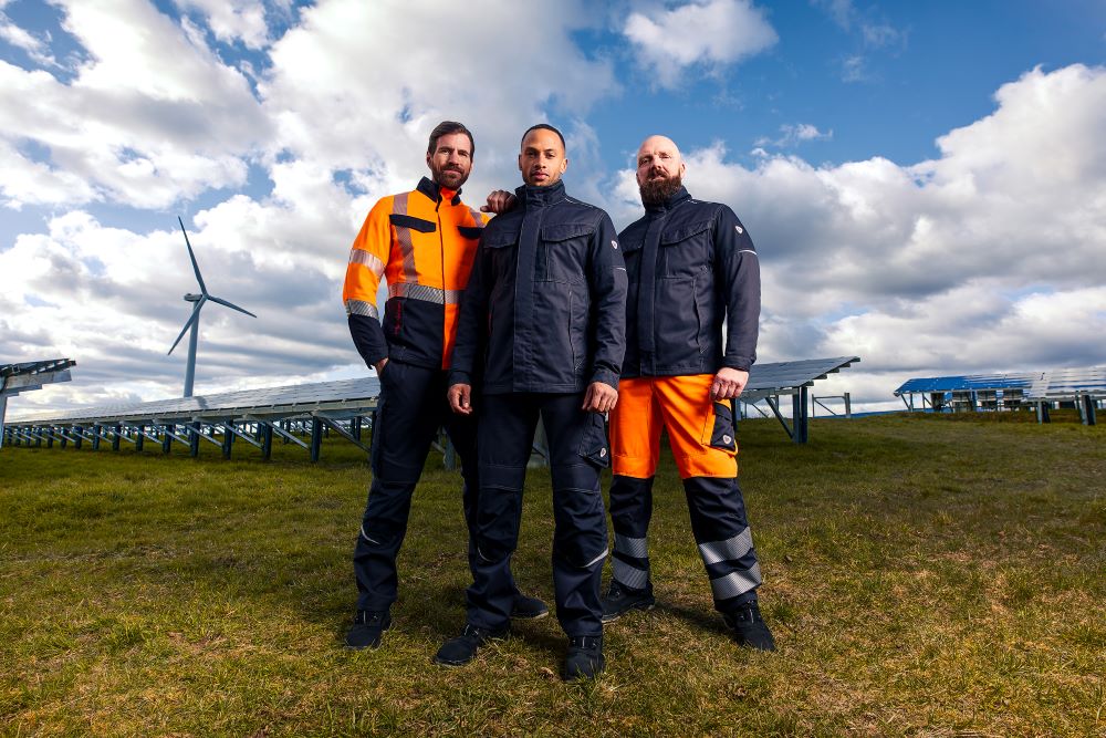 Drei Personen in moderner Arbeitskleidung vor Solar- und Windenergieanlagen.
