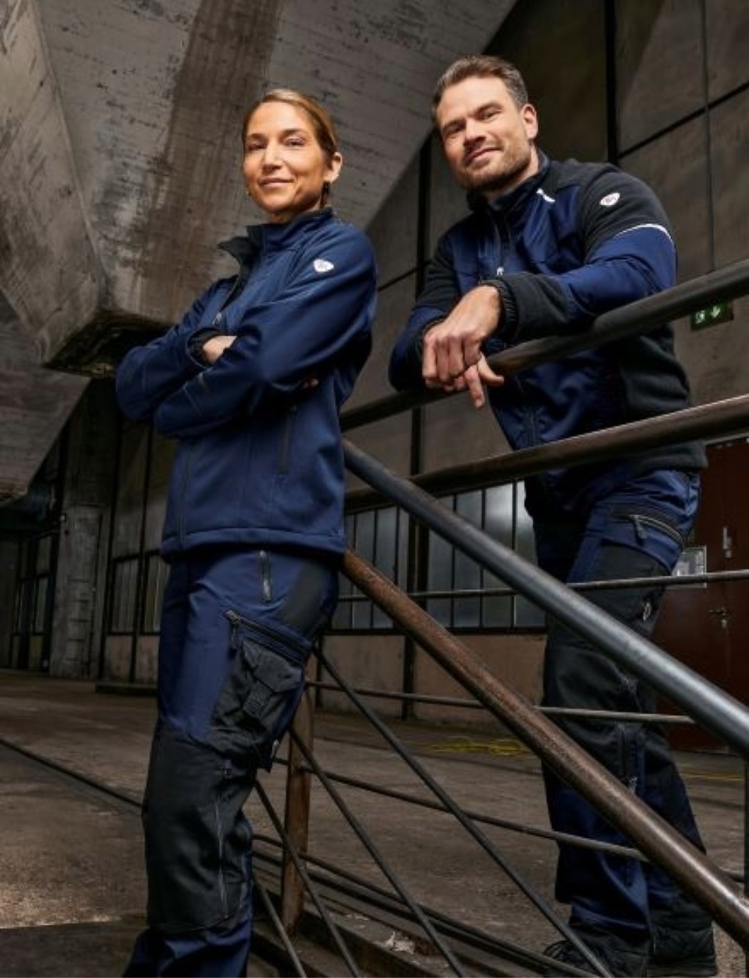 Frau und Mann in dunkelblauer Hybrid-Arbeitskleidung in altem Industriebau.