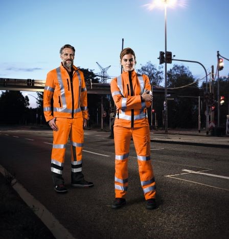 Een bouwvakker en een vrouwelijke bouwvakker staan 's nachts op straat in Hivi's werkkleding.