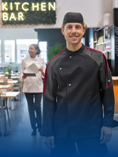 Ein Koch trägt Kochkleidung in anthrazit und posiert in einem Restaurant.