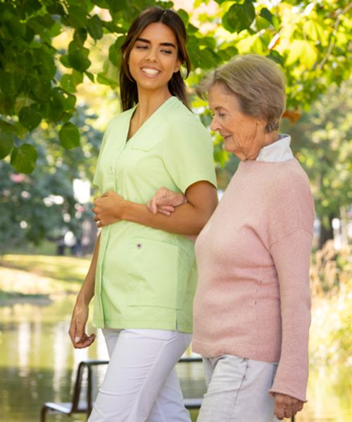 Alternpflegerin in grün-weißer Pflegekleidung geht mit Seniorin im Park spazieren.