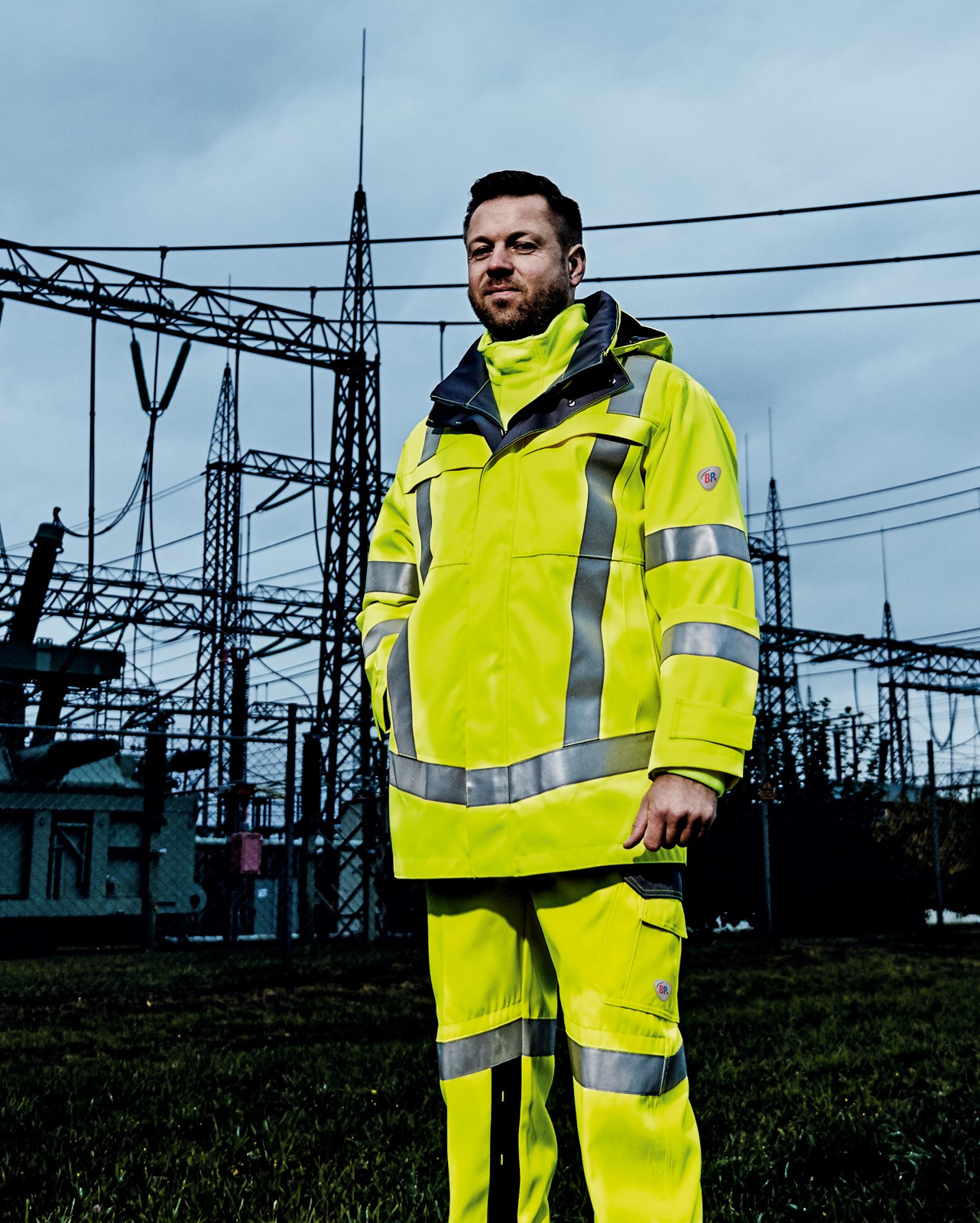 An energy manager wears neon yellow work clothes and stands in front of a substation.