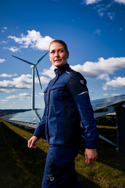 Eine Frau trägt Multinorm Schutzkleidung. Im Hintergrund ist eine Windkraftanlage und Solaranlagen zu sehen.