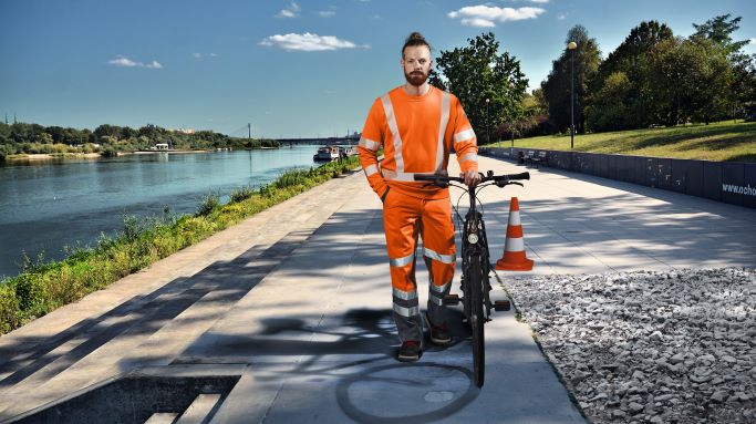 Mann in orangener Warnschutzkleidung schiebt Fahrrad am Rhein entlang.