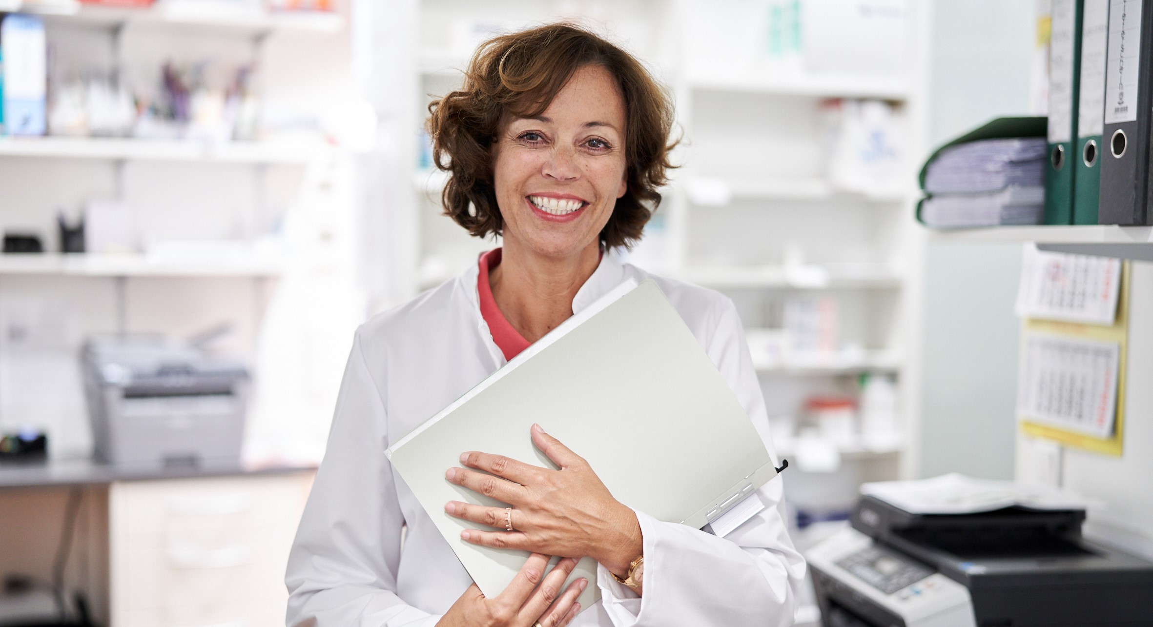 A pharmacist in white work clothes smiles at the camera.