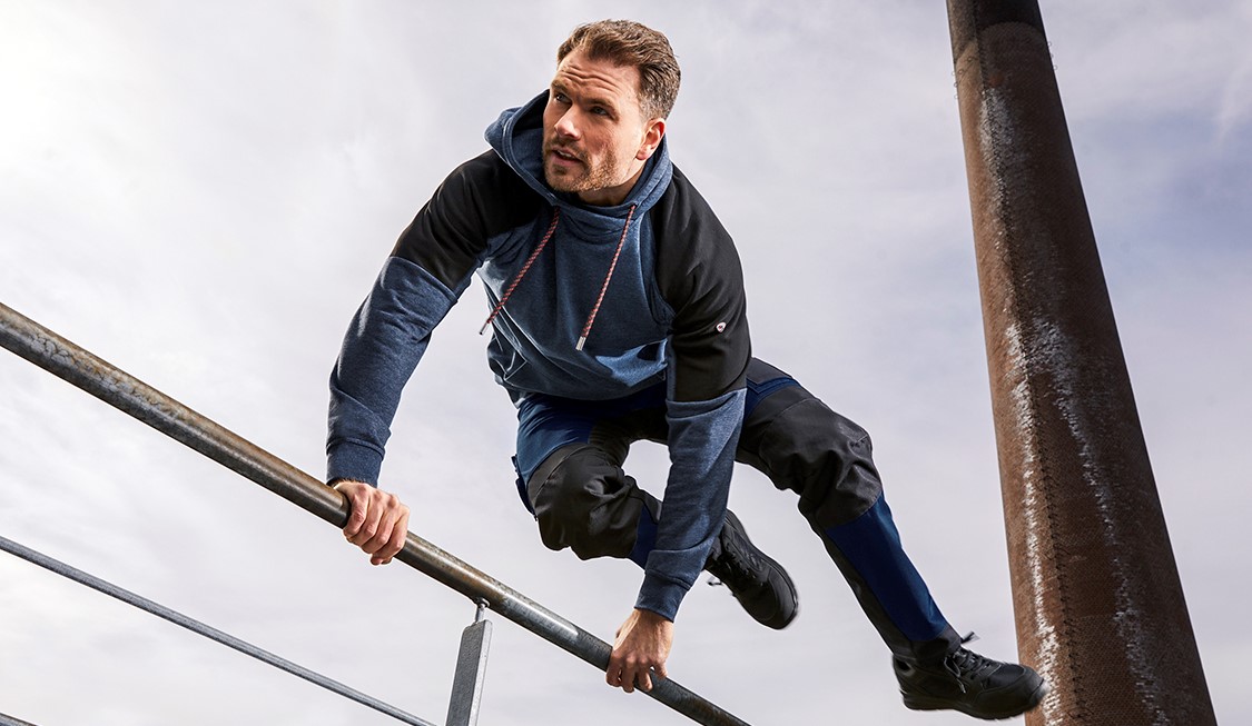 Ein Mann in einer hybriden und flexiblen Arbeitshose springt über eine Stange.