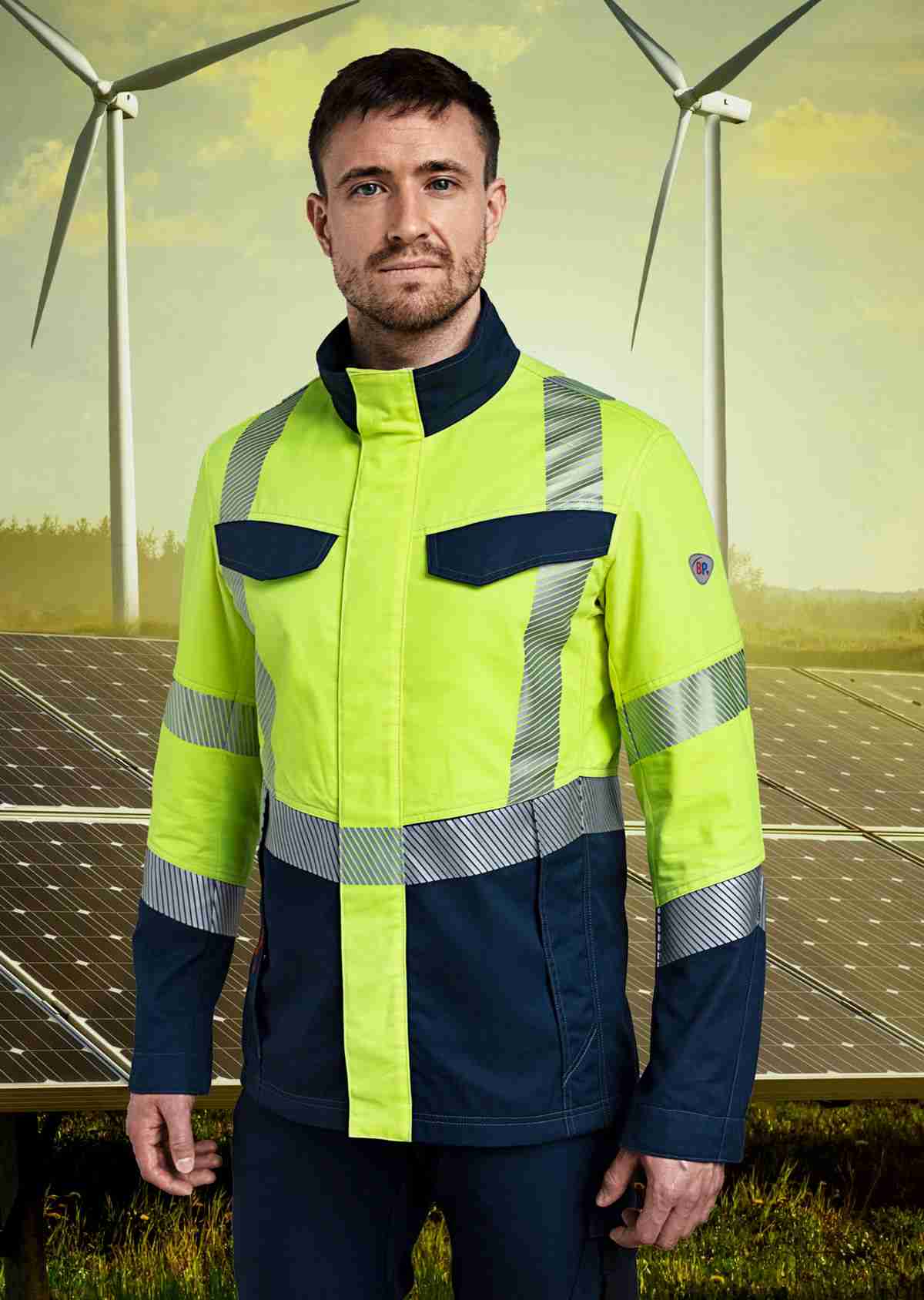 Ein Mann mit einer Multinorm Warnschutzjacke. Im Hintergrund sind Windkraftanlagen und Solaranlagen zu sehen.