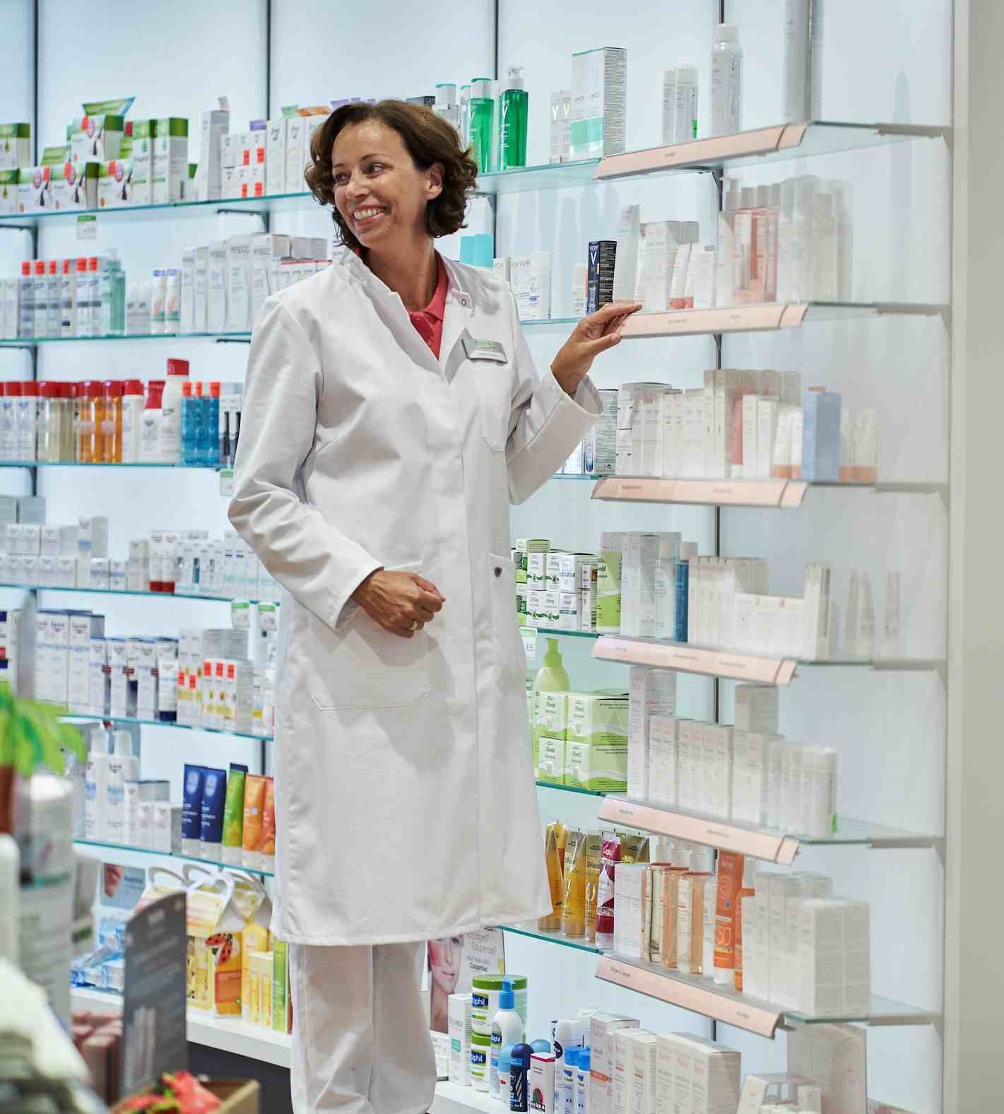 Eine Apothekerin in weißer Arbeitskleidung steht neben dem Medikamentenregal.