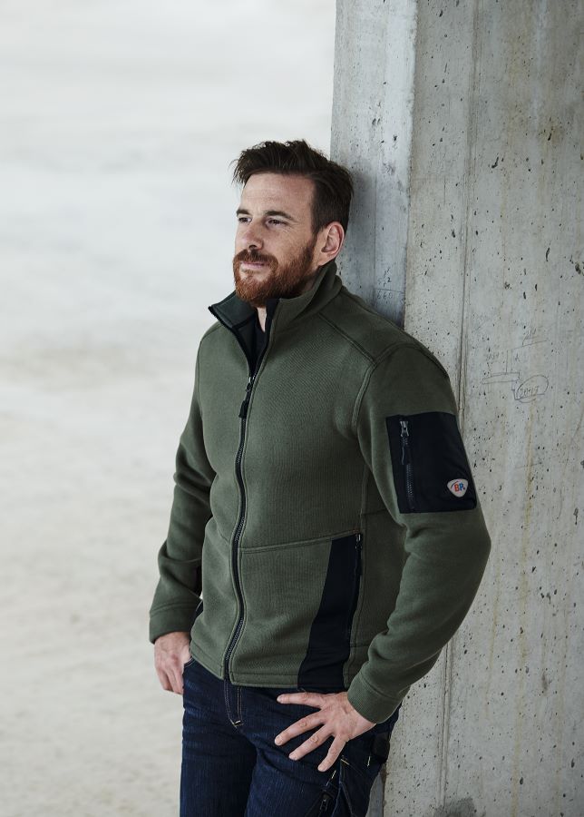 Handwerker in dunklegrüner Strick-Fleecejacke lehnt an eine Wand in einem Rohbau.