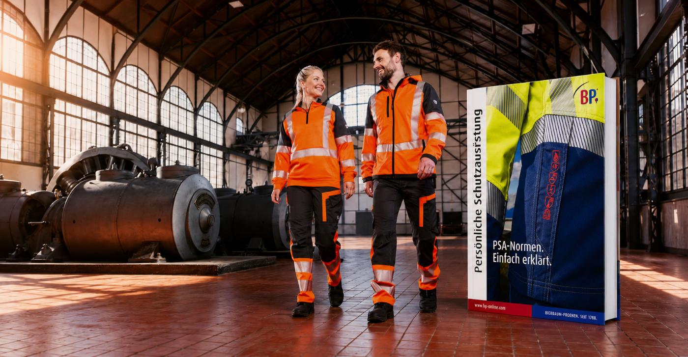  Eine Frau und ein Mann in Warnschutzkleidung laufen durch eine Industriehalle. Daneben ist das Buch "BP® Normenratgeber" abgebildet.