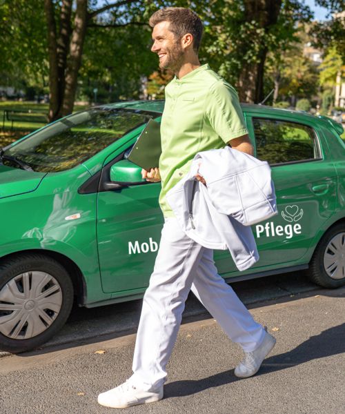 Krankenpfleger in grün-weißer Pflegekleidung steht vor seinem Pflegeauto.