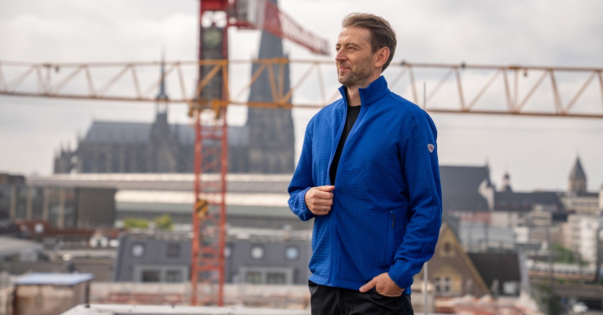 Mann in blauer Fleecejacke steht auf dem Dach, im Hintergrund der Kölner Dom.
