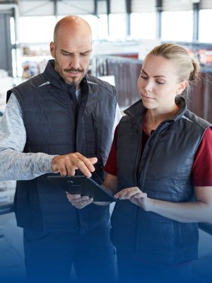 Ein Mann und eine Frau in blauen Arbeitswesten betrachten einen Arbeitsplan in einem Industriebetrieb.