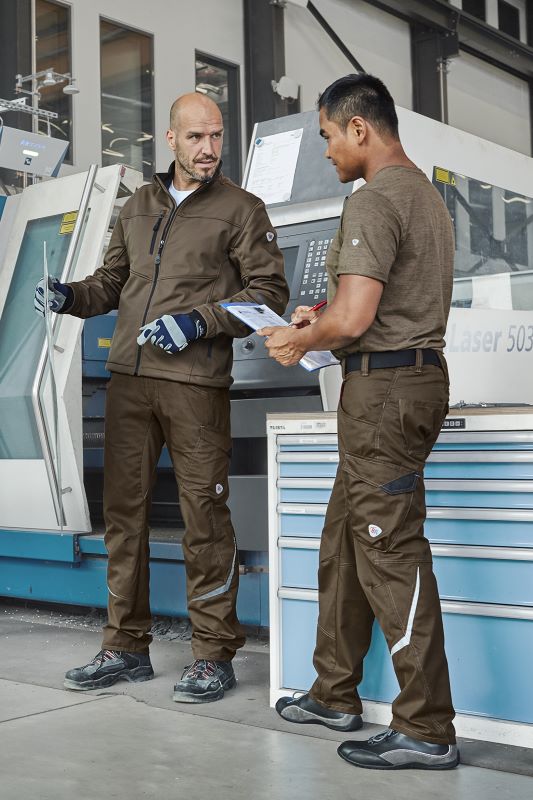 Zwei Facharbeiter in brauner Arbeitskleidung vor einer CNC-Anlage.