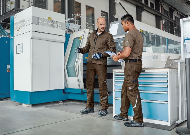 Zwei Arbeiter in grauer Arbeitskleidung arbeiten an CNC-Anlage.