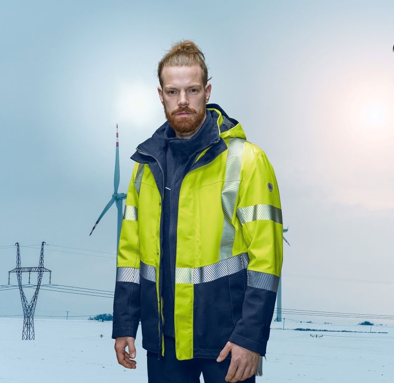 Een man draagt een weerbestendig jack volgens en343. Op de achtergrond zijn sneeuw en een windturbine te zien.