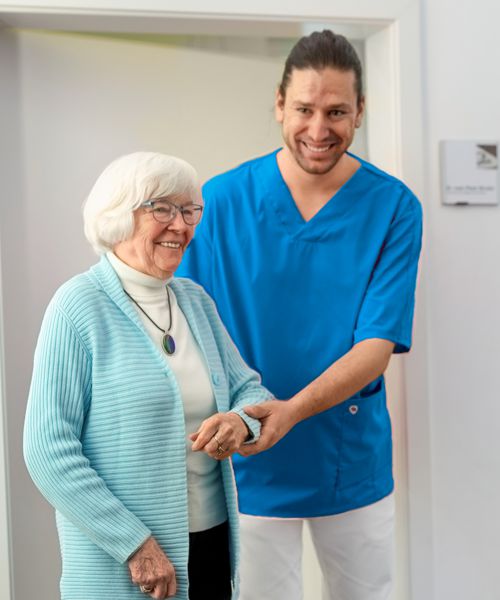 Carer in light blue coat helps senior citizen to walk.
