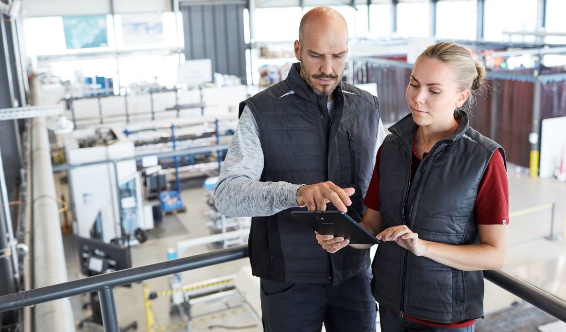 Ein Mann und eine Frau in blauen Arbeitswesten betrachten einen Arbeitsplan in einem Industriebetrieb.