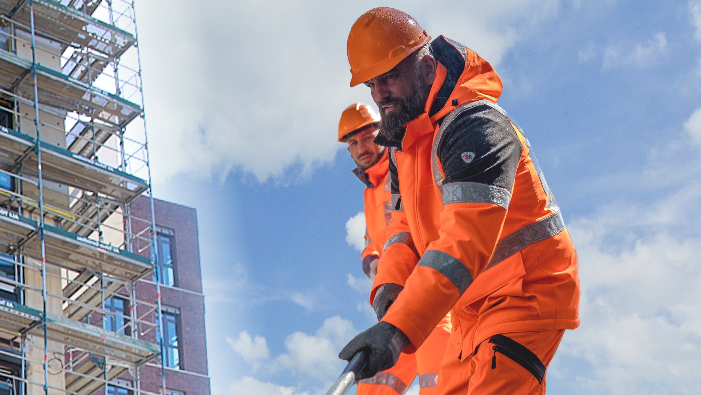 Two construction workers in orange high-visibility clothing are at work.