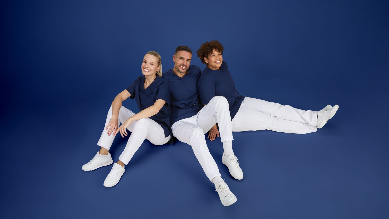 2 Pflegerinnen und 1 Pfleger in blau-weißer Pflegekleidung sitzen auf dem Boden.