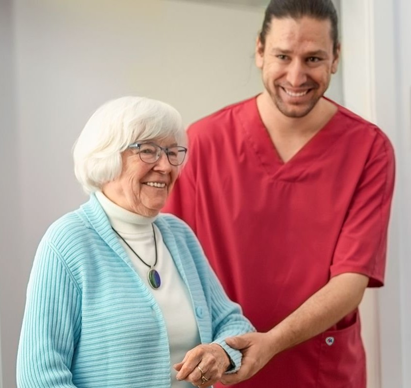 Altenpfleger in rotem Kasack hilft Seniorin beim Gehen.