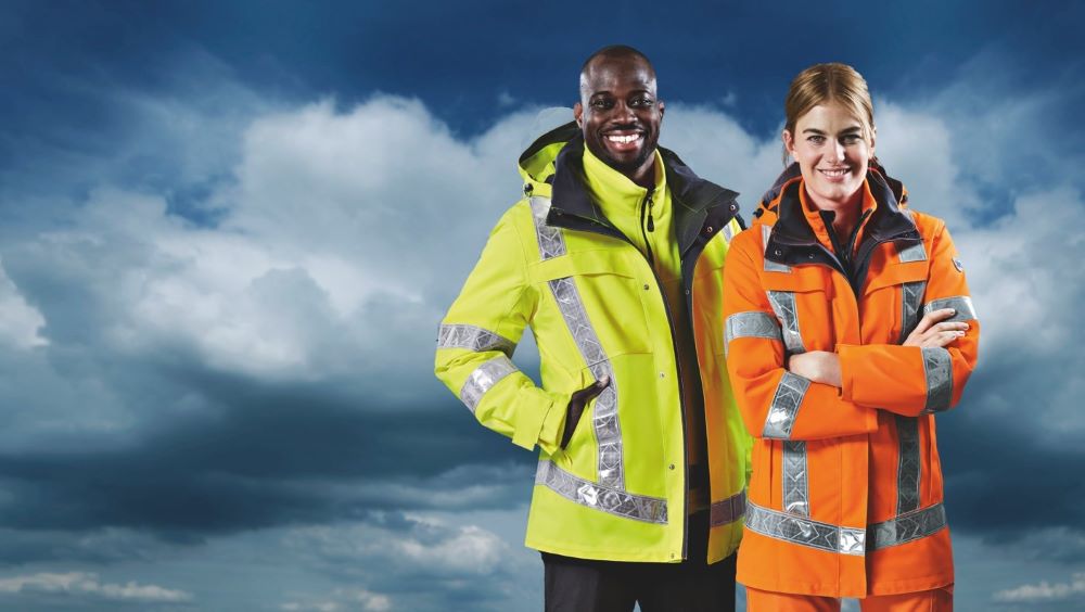 Zwei Personen in wetterfester Warnschutzkleidung vor dunklem Himmel, eine in Gelb, eine in Orange.