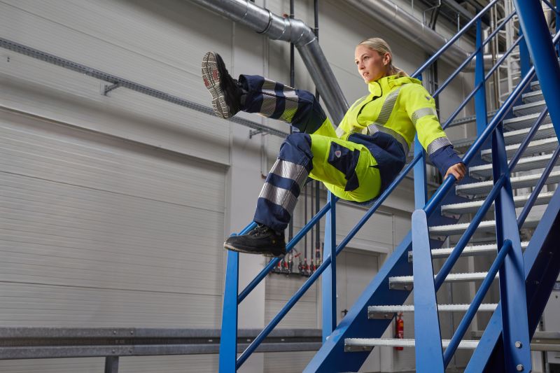 Frau in gelb-blauer Multinormkleidung springt Industrietreppe hinutnter.