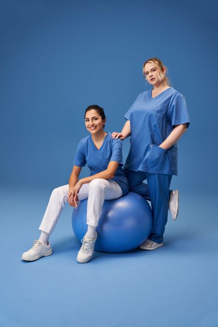 Zwei Pflegerinnen tragen azurfarbene Arbeitskleidung. Eine davon sitzt auf einem Gymnastikball.