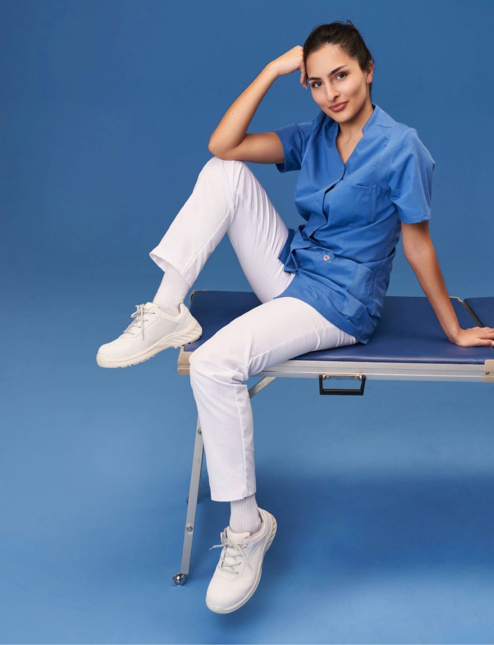 Une femme vêtue d'un pantalon de travail blanc et d'une casaque bleue à enfiler est assise sur une table de consultation.