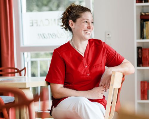 Une aide-soignante porte une casaque rouge et est assise sur une chaise, le sourire aux lèvres.